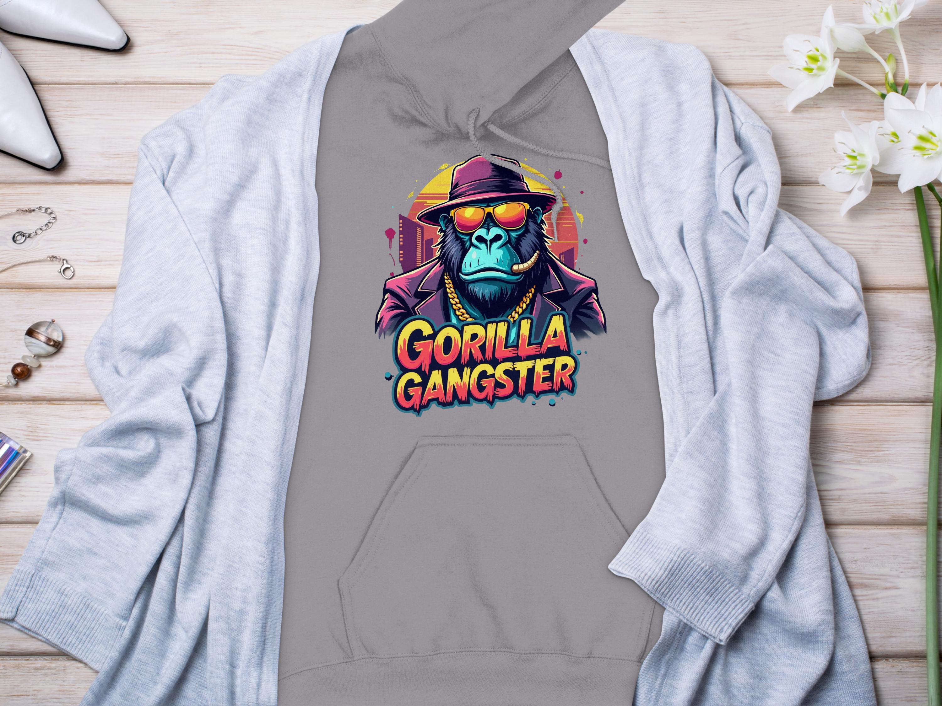 Garment Graphics presents a gray hoodie with vibrant gorilla artwork, featuring a cartoon gorilla in a fedora, sunglasses, and a gold chain with Gorilla Gangster in colorful letters. This streetwear piece is displayed on wood next to a gray cardigan, white shoes, and decorative items.