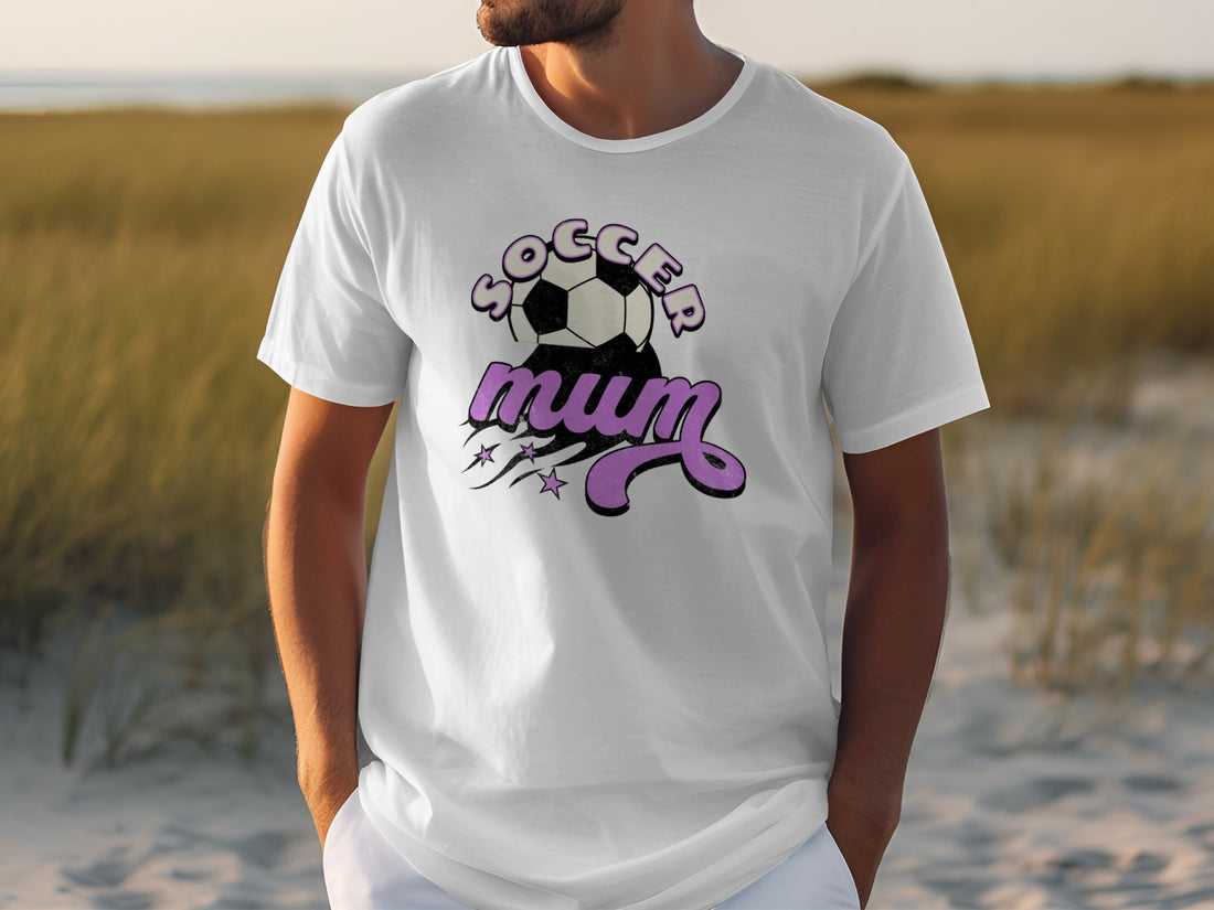 On a sandy path with grass behind, a person wears a classic fit T-shirt by Garment Graphics featuring SOCCER mum in black and pink letters, complete with a soccer ball and stars design.