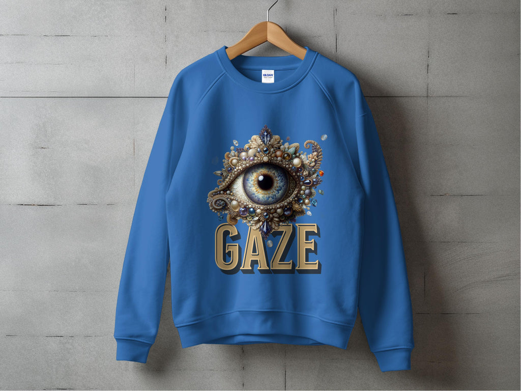 A blue unisex sweatshirt by Garment Graphics hangs on a wooden hanger, featuring an ornate eye design with pearls and jewels above GAZE in bold gold letters, set against a plain concrete wall.
