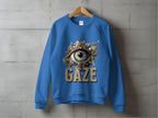 A blue unisex sweatshirt by Garment Graphics hangs on a wooden hanger, featuring an ornate eye design with pearls and jewels above GAZE in bold gold letters, set against a plain concrete wall.