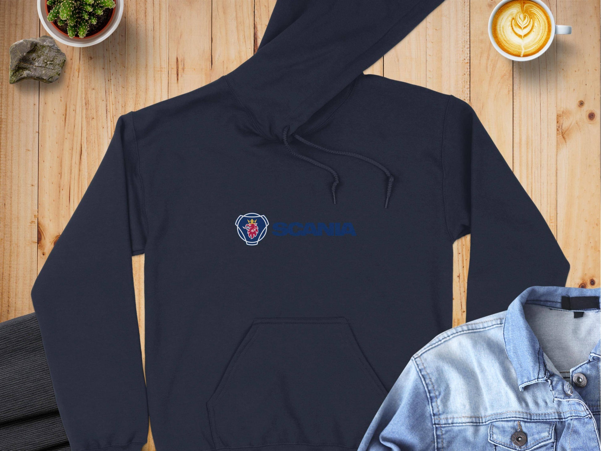 A Garment Graphics black Scania Hoodie with the blue “SCANIA” logo and emblem is laid on a wooden surface. Nearby, a denim jacket, a latte art coffee cup, a small plant, and folded dark pants add unique fashion elements to the scene.