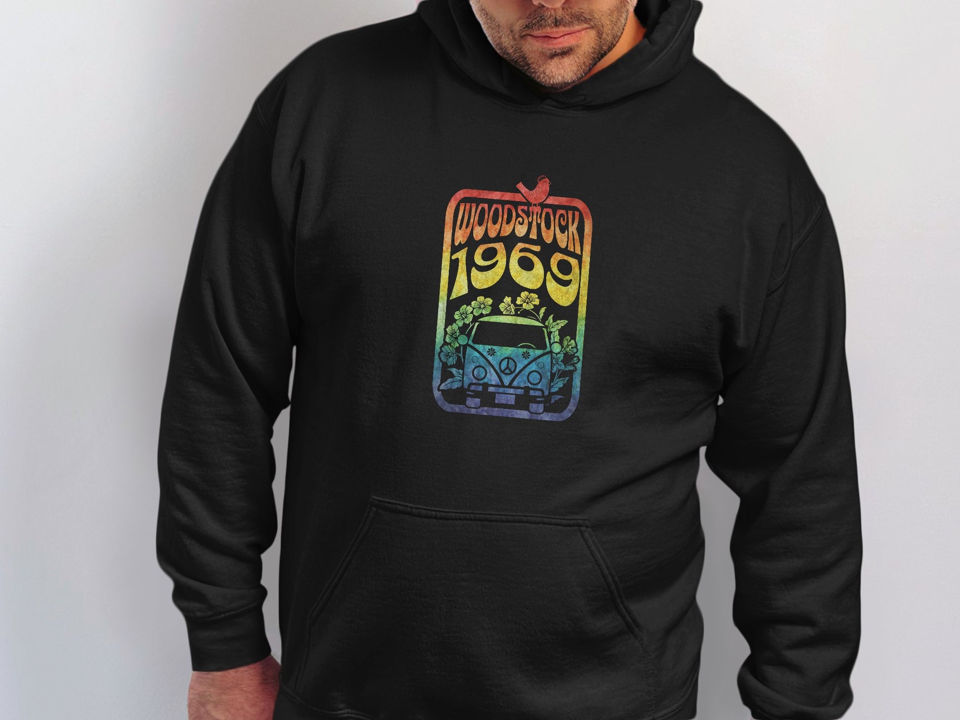 A person dons a retro black hoodie by Garment Graphics featuring a vintage Woodstock 1969 print. The design has a gradient with a car, flowers, and bird, against a plain white backdrop to emphasize its classic style.