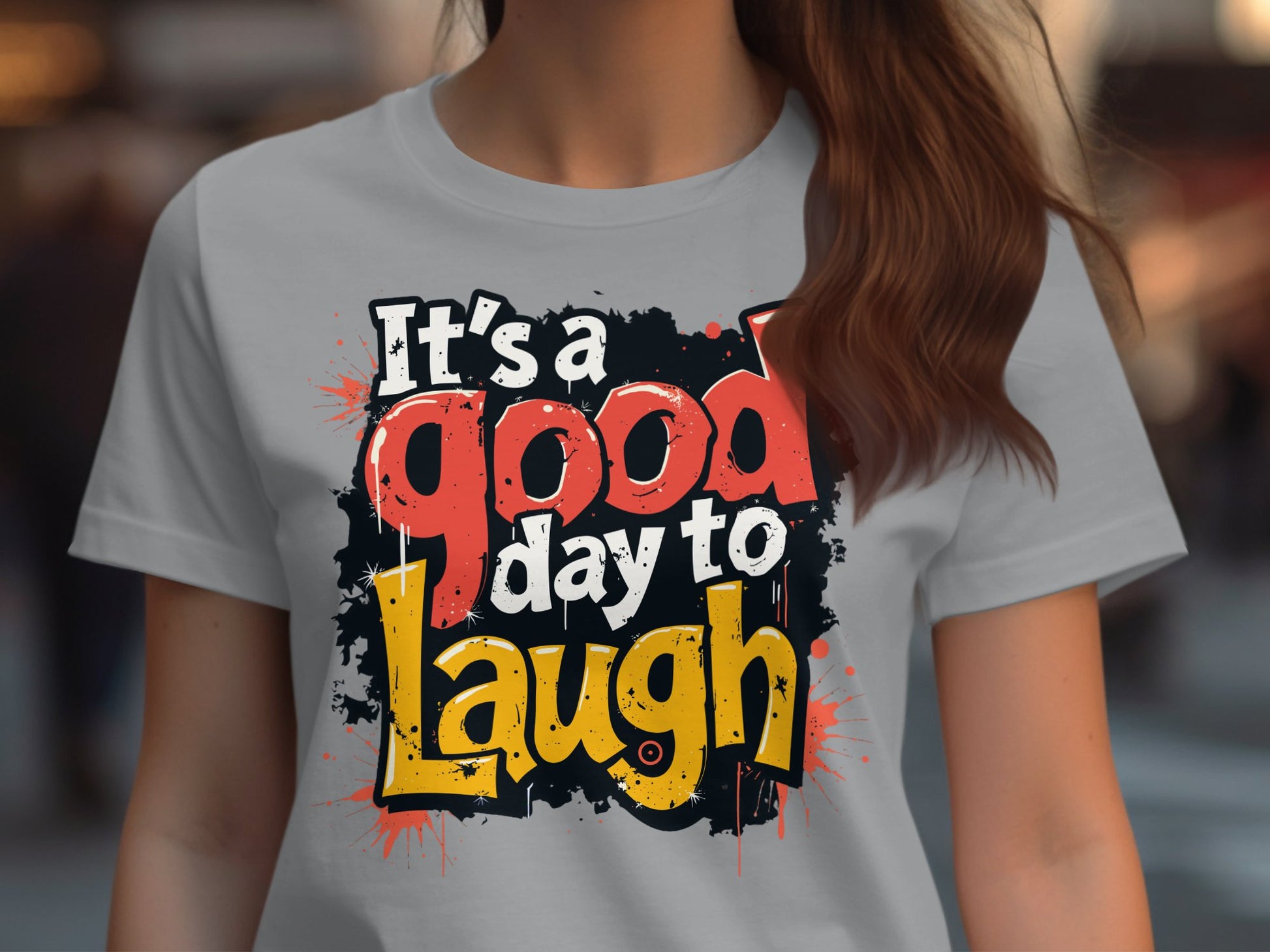 A person wearing a semi-fitted, light gray Garment Graphics T-shirt featuring bold red, orange, and yellow text, Its a good day to laugh, over a black splatter background. Their long brown hair enhances the positive vibe.