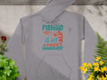 The Garment Graphics gray hoodie is essential for anglers, featuring Fishing Is My Anger Management in turquoise and orange. Adorned with a fish and lure, it’s stylishly displayed on a stone surface with plants enhancing its look.