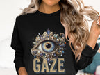 A person with long wavy hair wears Garment Graphics black graphic sweatshirt, featuring an ornate eye encircled by jewels and the word GAZE below. This unisex piece is ideal for those who love adding flair to their wardrobe.