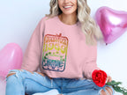 A woman sits cross-legged, smiling widely in a Garment Graphics vintage 1969 pink Woodstock festival sweatshirt. With a red rose in hand and heart-shaped pink balloons around her, she completes the retro vibe with ripped jeans.