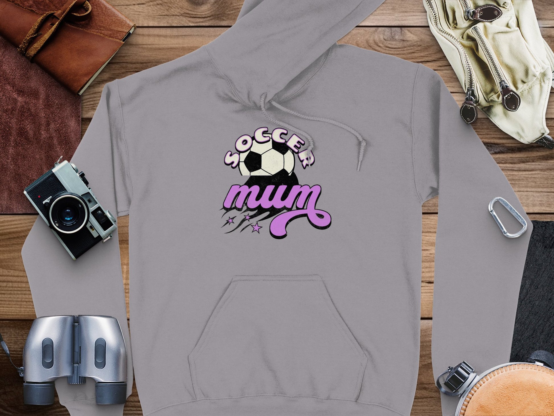 The Garment Graphics gray hoodie features Soccer Mum text, a soccer ball, and stars. Its cozy classic fit is displayed on a wooden surface with items like a vintage camera, leather book, binoculars, goggles, and a bracelet for an artful touch.