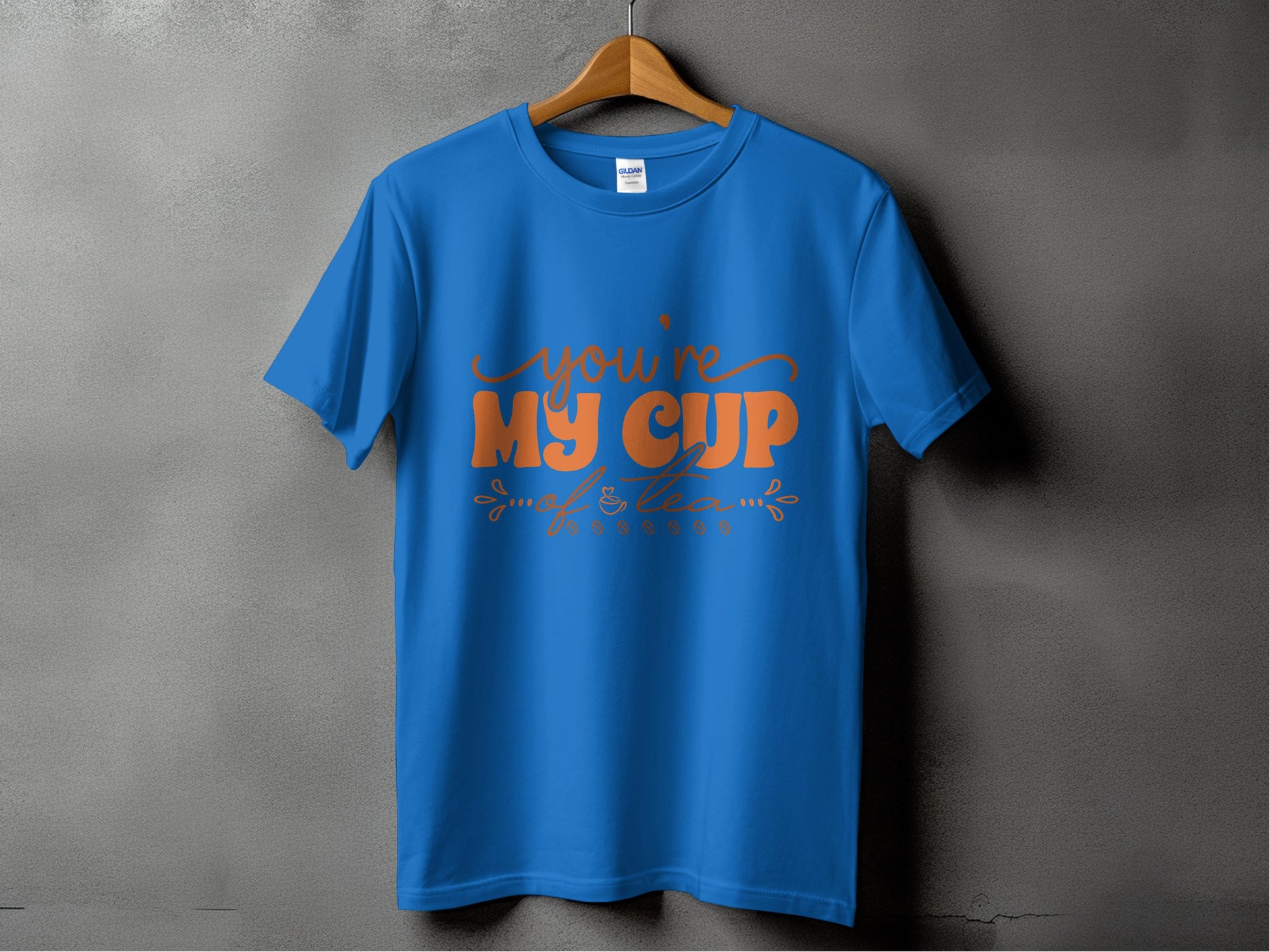 Against a gray backdrop, a blue 100% cotton T-shirt by Garment Graphics hangs on a wooden hanger. The front features stylized orange text saying, Youre My Cup of Tea, with decorative designs.