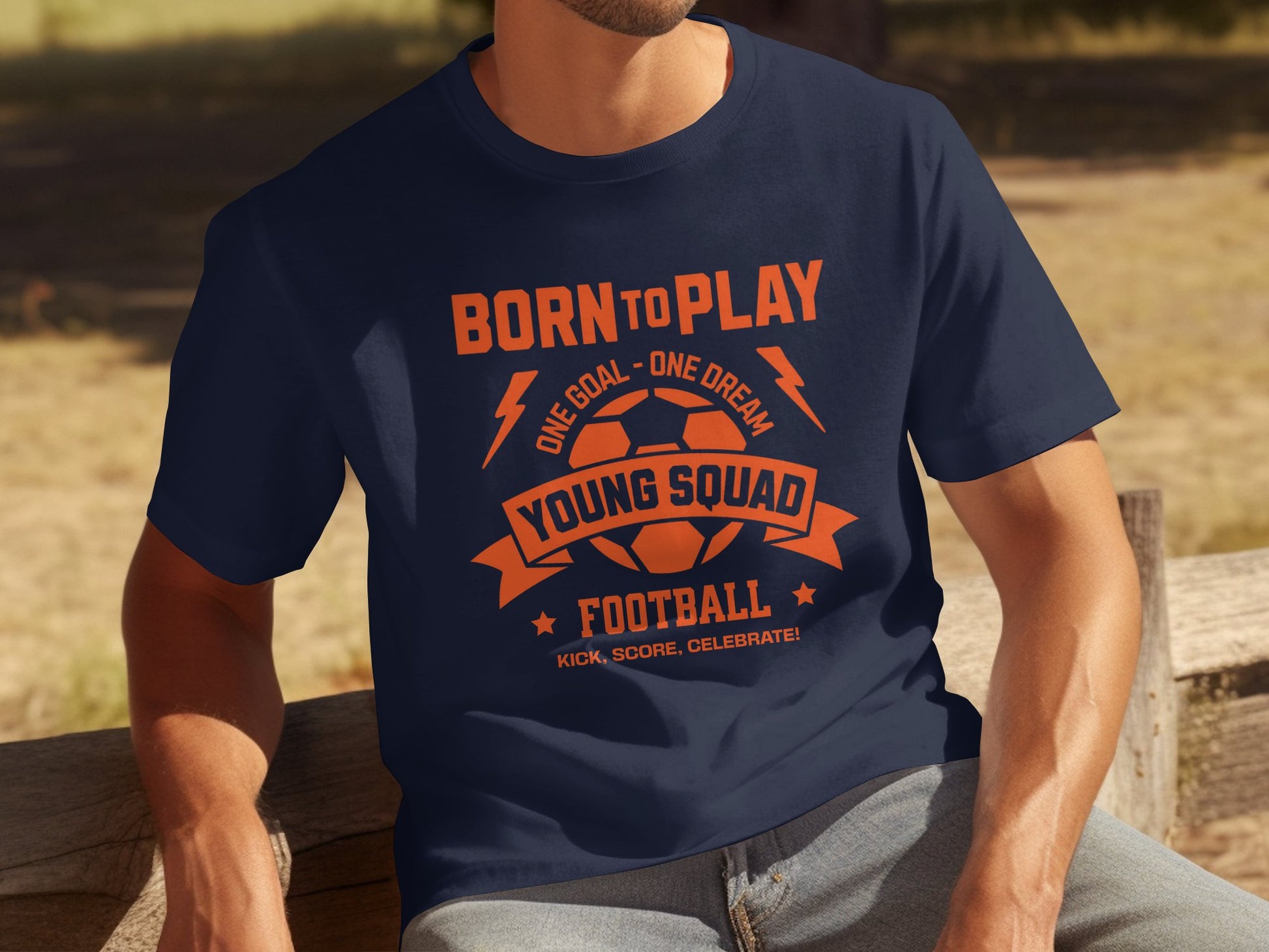 A person sits on a wooden bench in a navy blue Garment Graphics football T-shirt featuring the phrase BORN TO PLAY, ONE GOAL - ONE DREAM, YOUNG SQUAD, with a soccer ball graphic prominently displayed.