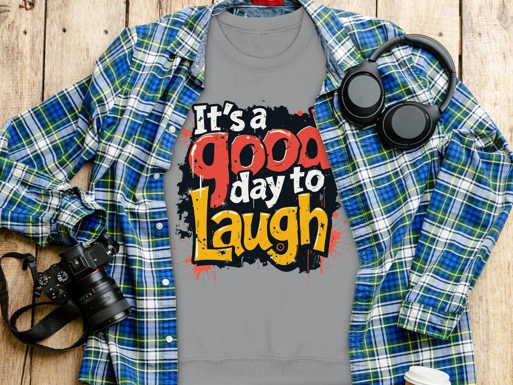 The Garment Graphics gray T-shirt, featuring bold, colorful Its a good day to laugh text and medium-heavy fabric in unisex sizing, is displayed on a wooden surface with a blue and green plaid shirt, headphones, a camera, and a white coffee cup.