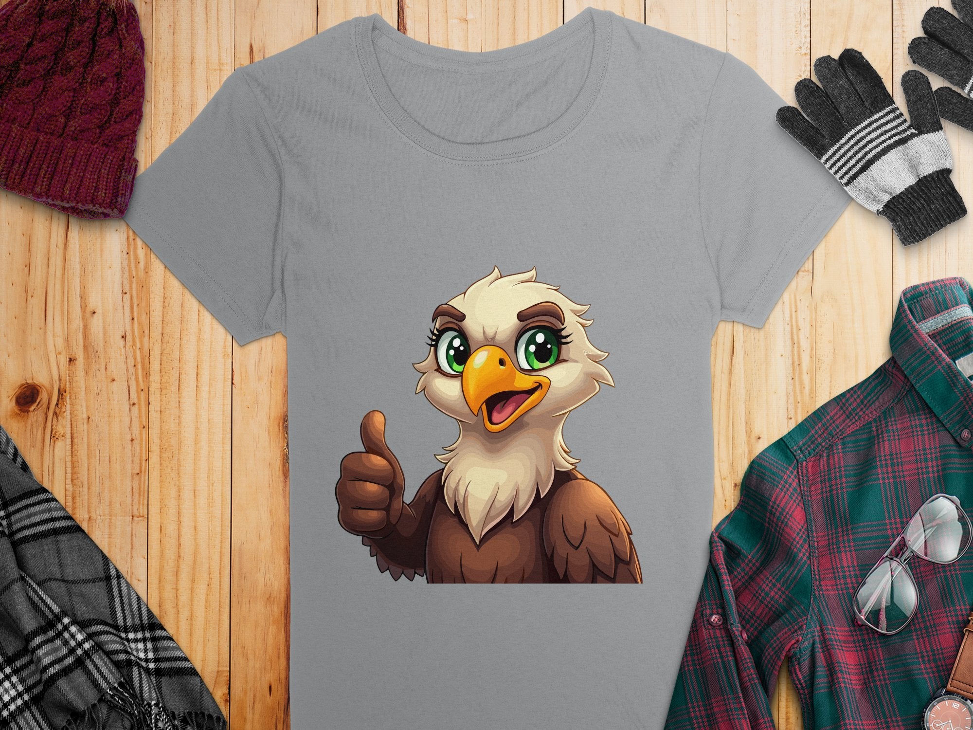A Garment Graphics grey cartoon T-shirt featuring a thumbs-up eagle lies on a wooden surface, ideal for animal lovers. Nearby are a red knit beanie, black-and-white gloves, a green plaid shirt, and a black-and-white striped scarf.