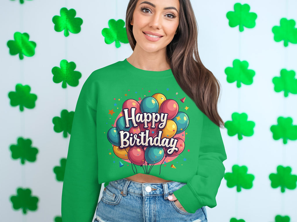 A woman stands by a wall of green shamrocks, wearing a Garment Graphics sweatshirt with colorful balloons and Happy Birthday text, ideal for celebrations. Her long hair and denim shorts complete the festive look with unisex sizing considered.