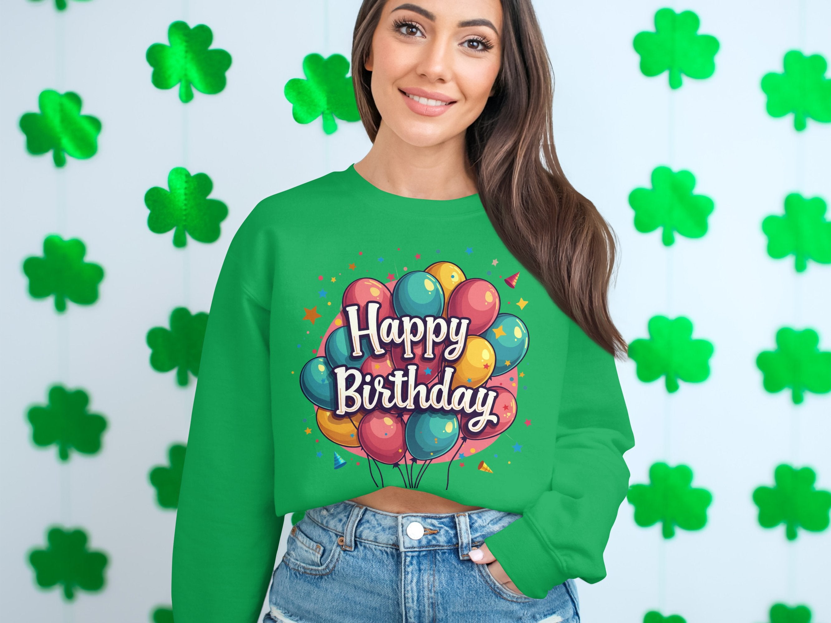 A woman stands by a wall of green shamrocks, wearing a Garment Graphics sweatshirt with colorful balloons and Happy Birthday text, ideal for celebrations. Her long hair and denim shorts complete the festive look with unisex sizing considered.
