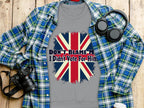A Garment Graphics gray t-shirt featuring the Union Jack and text Dont Blame Me, I Didnt Vote For Him lies on a wooden surface with a blue flannel shirt, a camera, headphones, and a cup nearby.