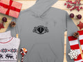 A gray Scania Power Eight Hoodie by Garment Graphics, showcasing a bold chest design with stars and stylized elements, lies next to a classic fit knitted sweater among red and white wrapped gifts, pine cones, berries, and decorative stars on a wooden surface.