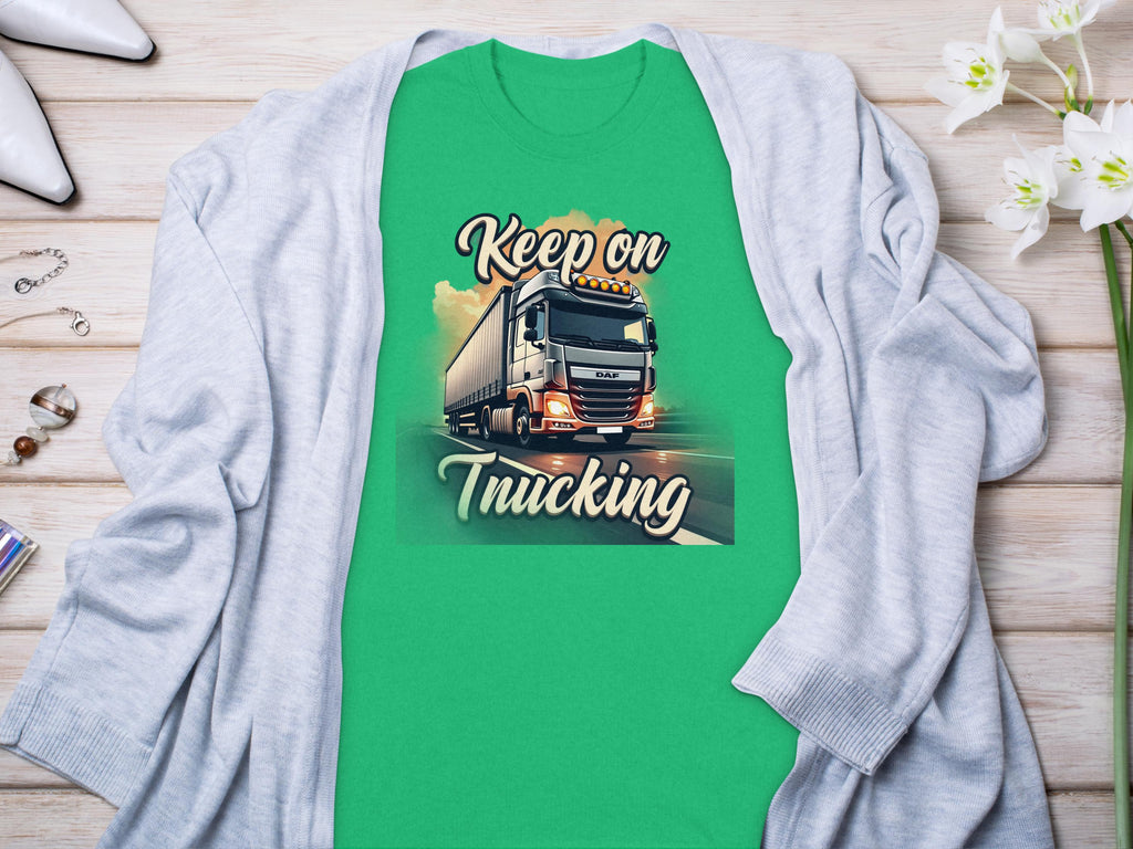 A green Garment Graphics t-shirt with a truck graphic and Keep on Trucking text lies on wood. Its ideal for truck fans, surrounded by a gray cardigan adorned with white flowers, and jewelry scattered artfully nearby.