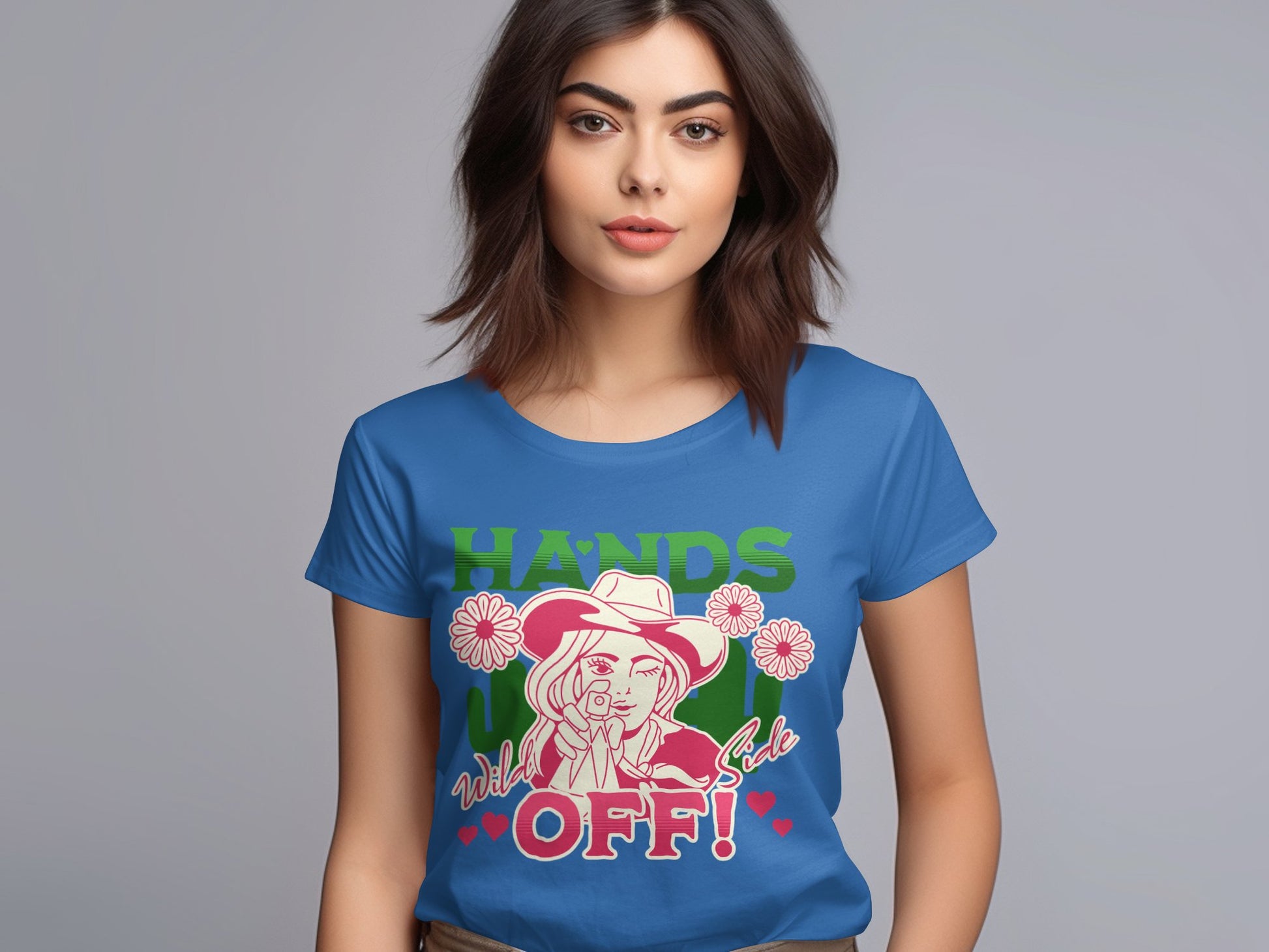 A brown-haired woman dons a blue Garment Graphics T-shirt with a bold design, featuring a person in a hat surrounded by flowers and the vibrant text HANDS OFF! in various colors, showcasing her unique style.