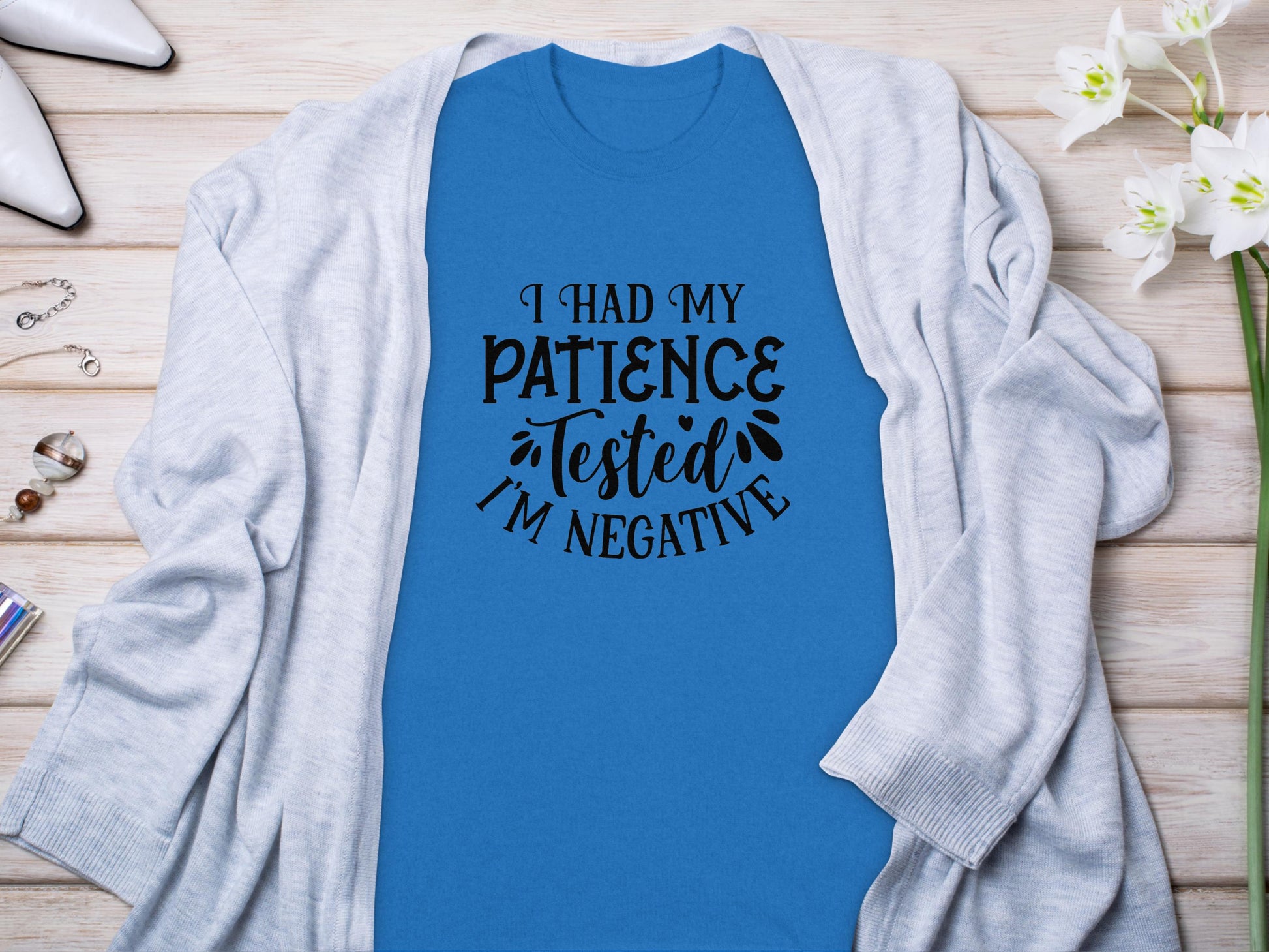 A Garment Graphics blue T-shirt, featuring the phrase I Had My Patience Tested Im Negative in black text, conveys sarcasm. Its styled on a wooden surface with a light gray cardigan, alongside white shoes and a small white floral arrangement to accentuate its humor.