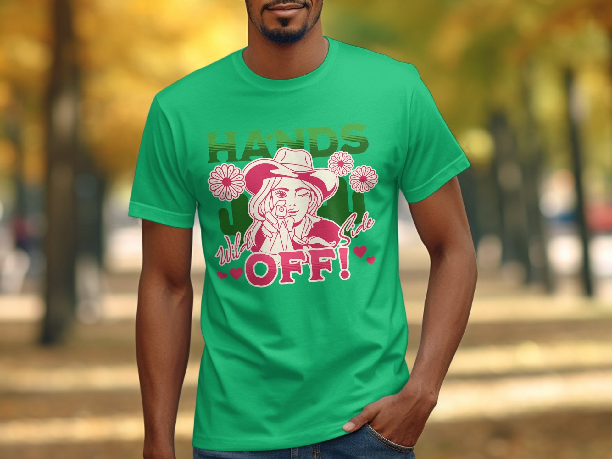 A man wears Garment Graphics bold green t-shirt featuring a graphic of a hatted person with flowers, striking HANDS OFF! text, and smaller wild and safe letters. This classic fit shirt stands out against the parks trees and blurred paths.