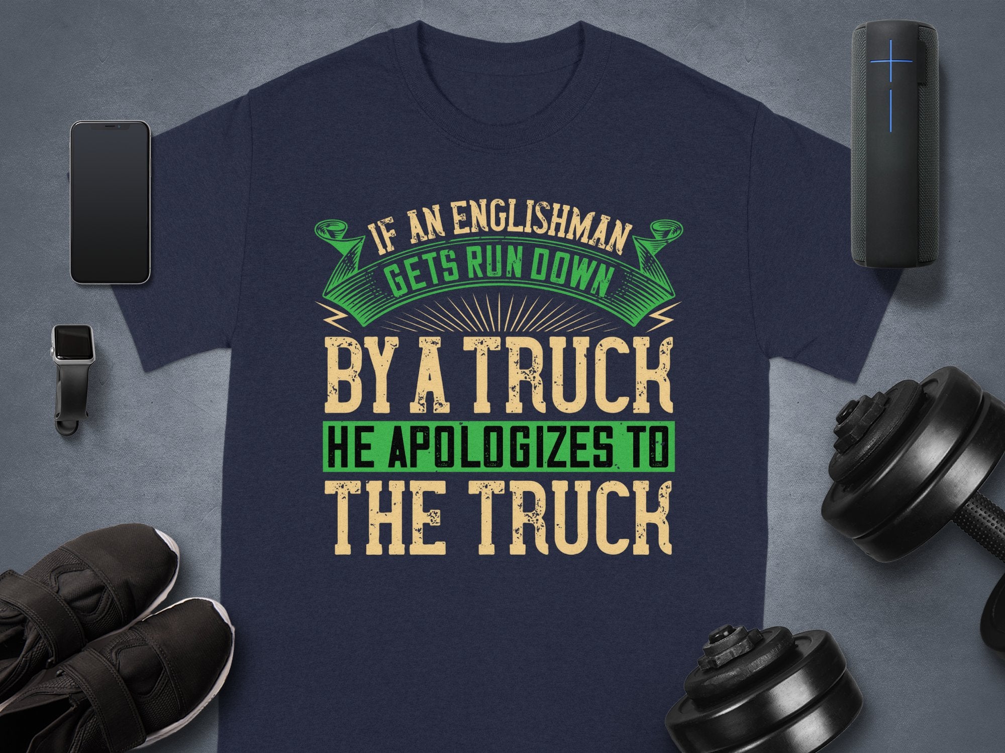 A flat lay image features Garment Graphics navy t-shirt with the witty If an Englishman gets run down by a truck, he apologizes to the truck in green and tan fonts. Nearby are a smartwatch, sneakers, speaker, and dumbbell. This humorous tee underscores English politeness.