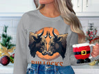 A person with long hair holds a Santa torso mug and wears an eye-catching gray Garment Graphics sweatshirt with two bulls facing each other, an orange circle behind them, and BULLOCKS below. The background reveals a decorated kitchen.