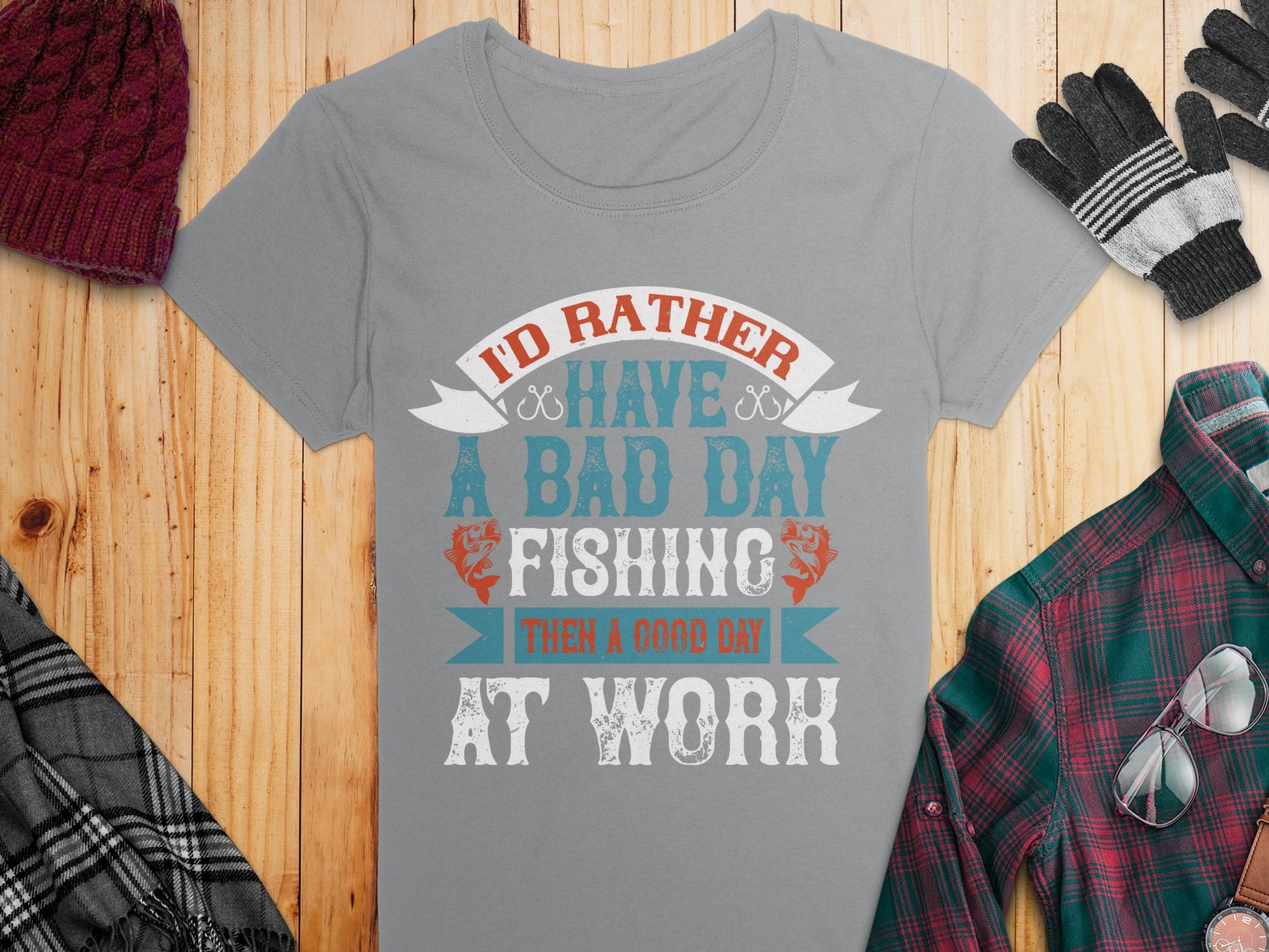 A Garment Graphics gray fishing T-shirt in womens sizing features Id rather have a bad day fishing than a good day at work in red, white, and blue. Perfect for enthusiasts, it’s styled with a plaid shirt, gray gloves, and knit hat on a wooden backdrop.