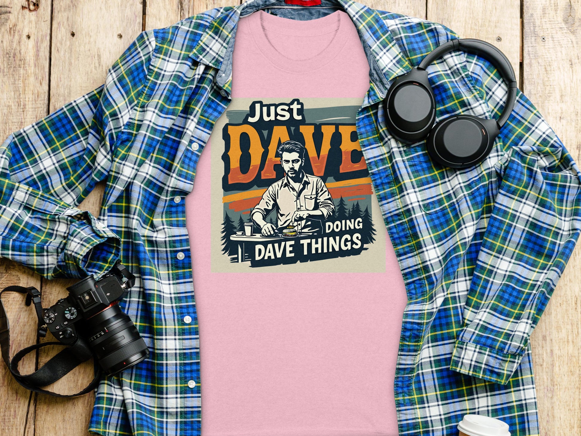 Garment Graphics classic fit pink shirt features the Just Dave Doing Dave Things graphic, depicts someone at a table. It pairs perfectly with a blue plaid shirt, headphones, camera, and coffee cup on a wooden surface for a relaxed vibe.