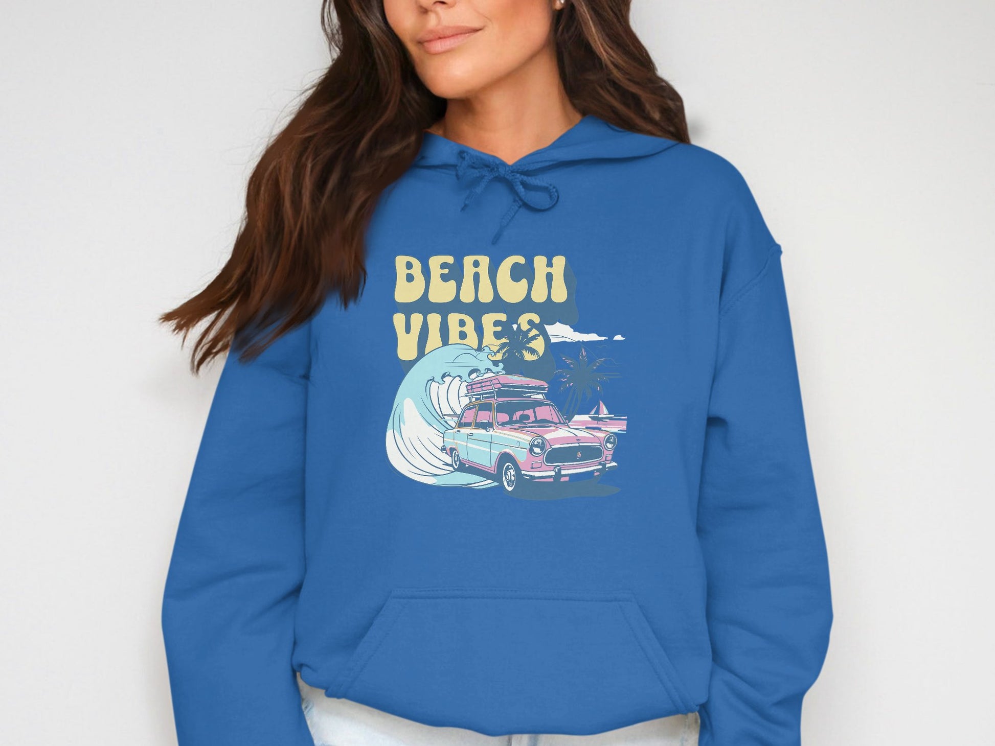 A woman in a vibrant blue hoodie by Garment Graphics, featuring BEACH VIBES text and an illustration of a pink vintage car navigating a large ocean wave with palm trees, showcasing retro beach style.