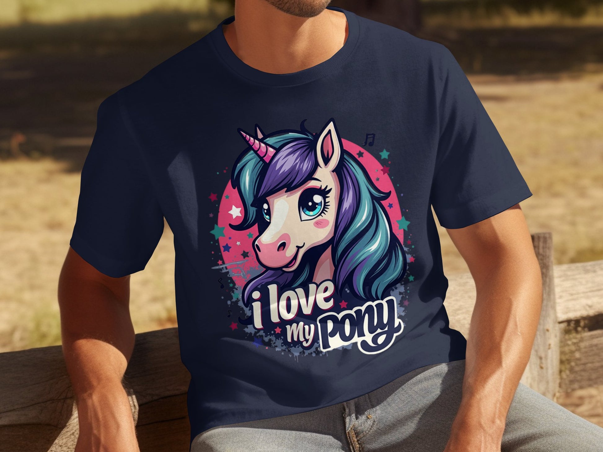 A unicorn enthusiast relaxes on a wooden bench, wearing Garment Graphics dark blue pony graphic T-shirt with a colorful unicorn illustration. The shirt reads I love my pony beneath a starry backdrop, showcasing their passion for these mythical creatures.