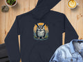 A black hoodie featuring a fierce owl design from Garment Graphics lies on a wooden surface, flanked by a denim jacket and latte art coffee for coziness. A small plant and rock add to the inviting ambiance.