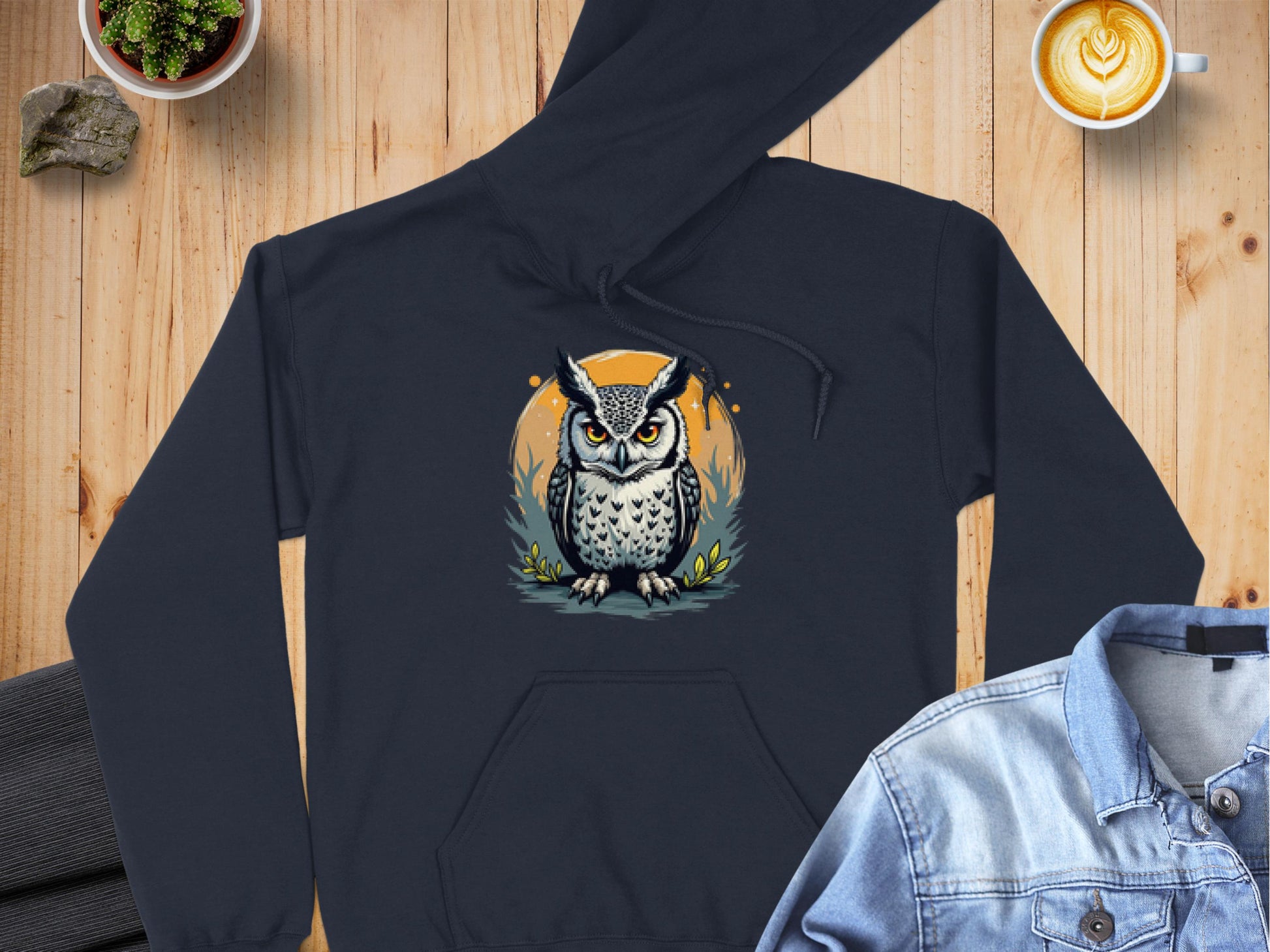 A black hoodie featuring a fierce owl design from Garment Graphics lies on a wooden surface, flanked by a denim jacket and latte art coffee for coziness. A small plant and rock add to the inviting ambiance.