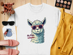 The Garment Graphics white No Prob-Llama sweatshirt, featuring a llama in red sunglasses, humorously complements a mustard yellow jacket and pink loafers. On a wooden surface with makeup items, extra sunglasses, brushes, a watch, and a blue pouch, this sweatshirt offers inclusive unisex sizing.