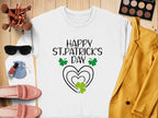 A festive white sweatshirt by Garment Graphics featuring Happy St. Patricks Day in black, with a heart and clover design, styled alongside a yellow blazer, pink shoes, sunglasses, makeup brushes, and a vibrant makeup palette on a wooden background.