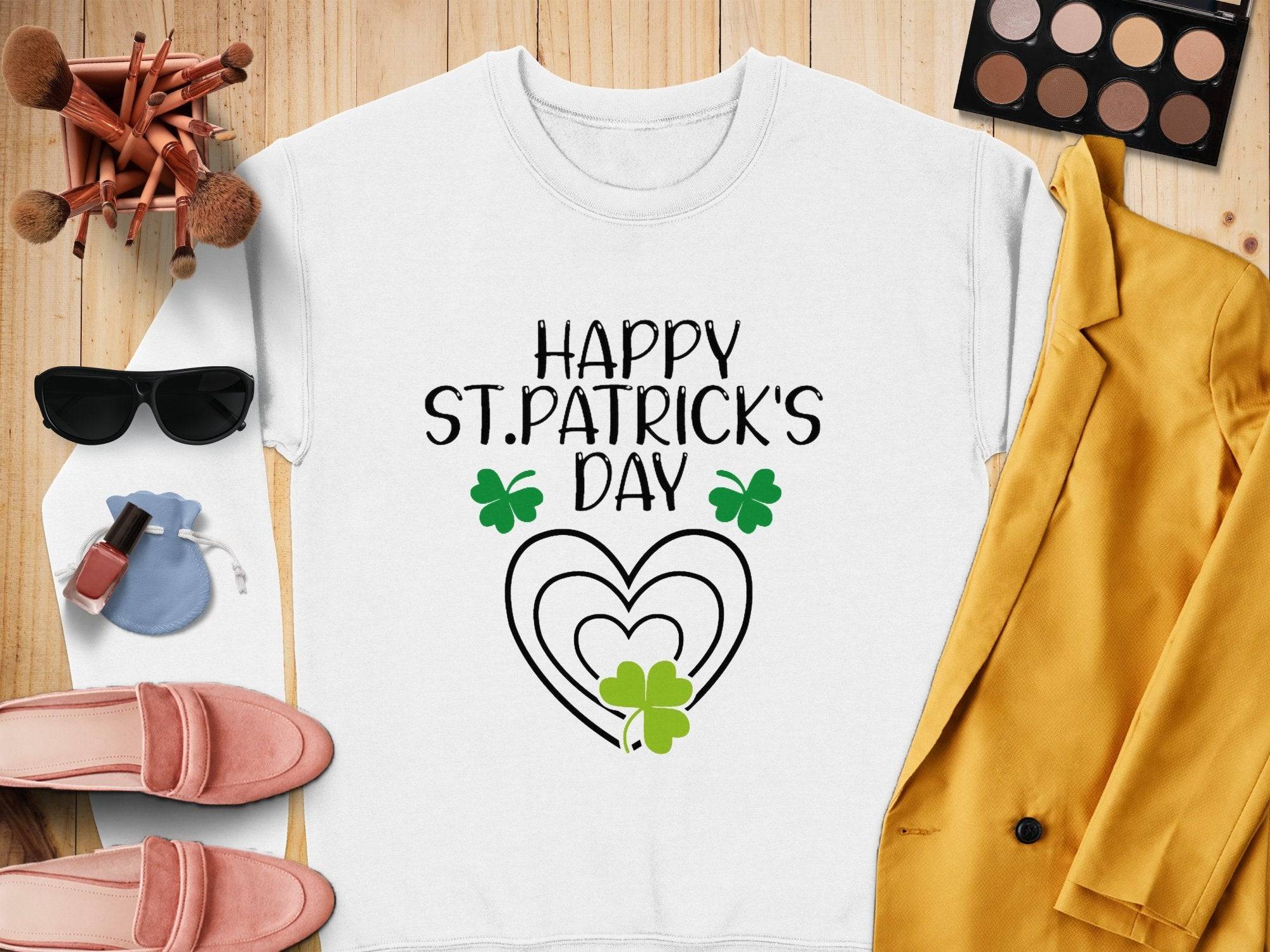 A festive white sweatshirt by Garment Graphics featuring Happy St. Patricks Day in black, with a heart and clover design, styled alongside a yellow blazer, pink shoes, sunglasses, makeup brushes, and a vibrant makeup palette on a wooden background.