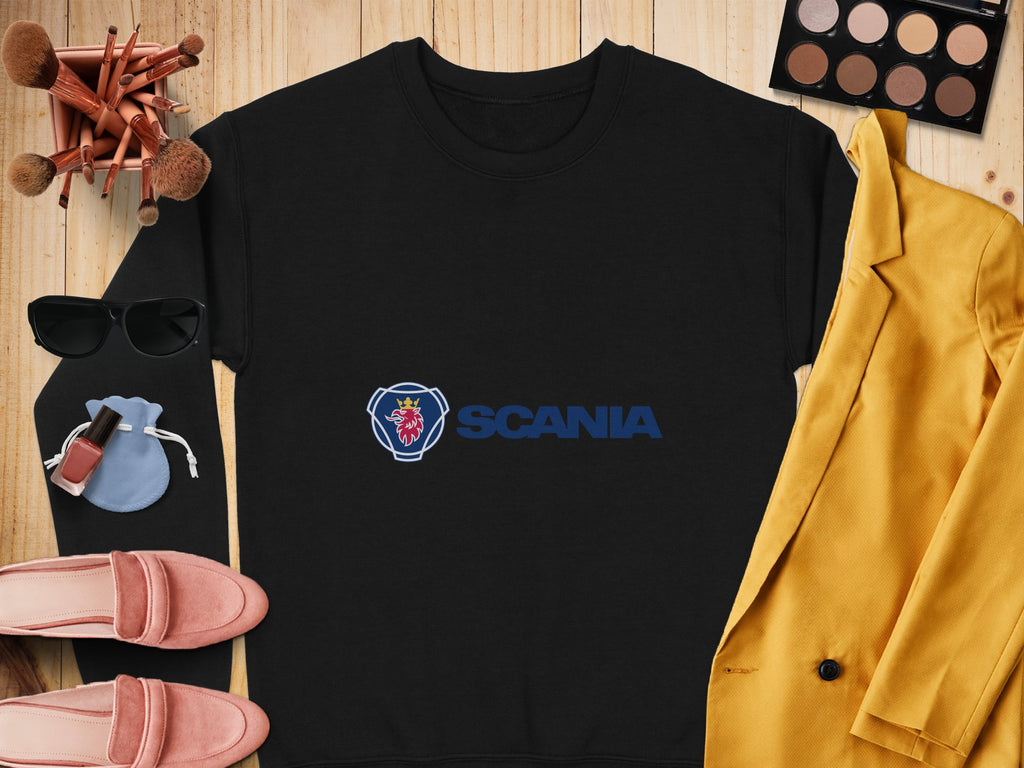 A black unisex sweater featuring the Scania logo, by Garment Graphics, is displayed on wood. It’s complemented by a yellow jacket, makeup items, sunglasses, pink shoes, a blue pouch, and a palette – an ideal ensemble for casual outings.