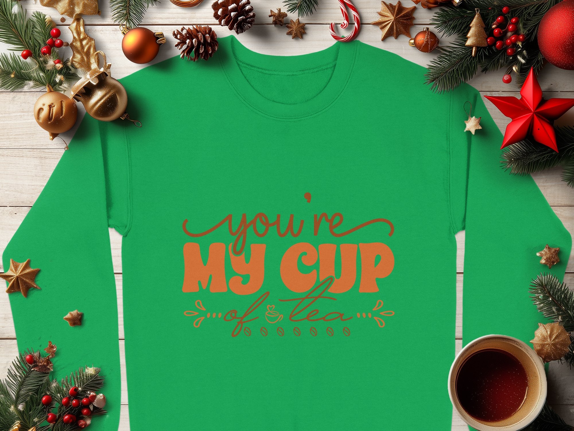 The Garment Graphics green shirt, perfect for tea enthusiasts, features Youre My Cup of Tea in orange and brown text. Set against holiday decorations—pine branches, pinecones, ornaments, and a cozy cup of tea—it rests on a wooden surface.