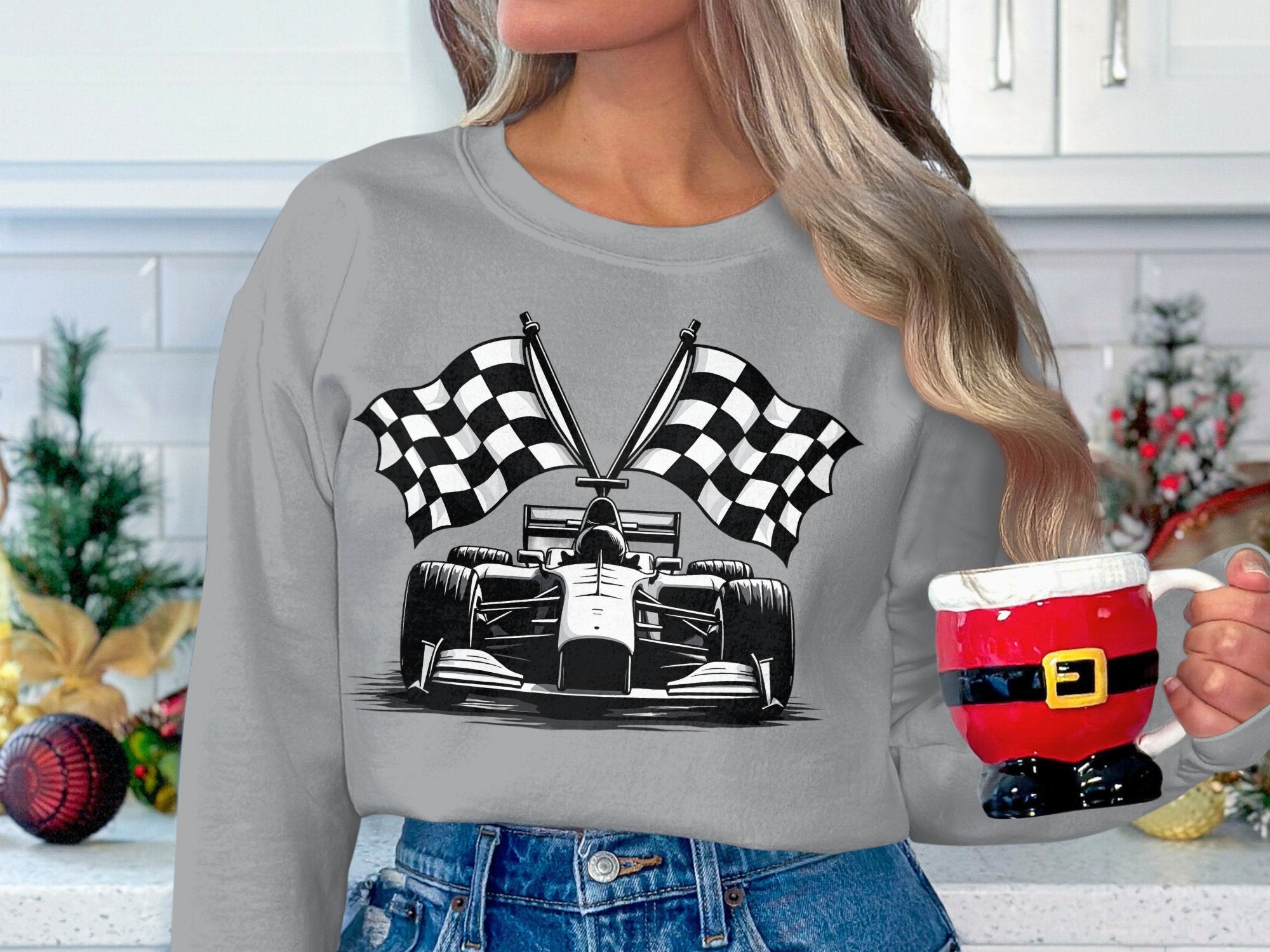 A long-haired person in a gray Garment Graphics racing car sweatshirt, featuring a bold checkered flag print, holds a Santa-themed mug. They stand in a kitchen with white cabinets and festive plants and ornaments around them.