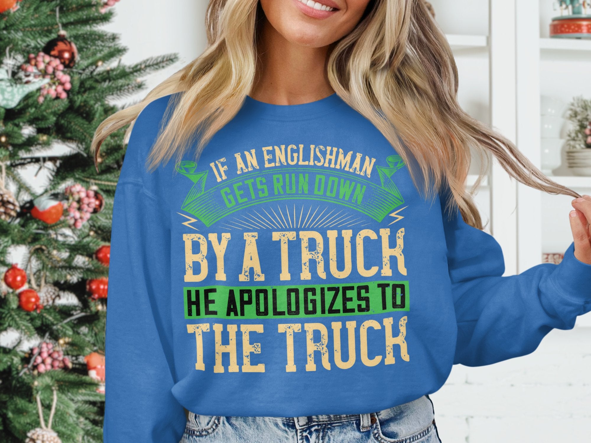 Wearing Garment Graphics blue sweatshirt featuring the bold, humorous design: If an Englishman gets run down by a truck, he apologizes to the truck, a person stands before a festively decorated Christmas tree.