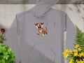 The gray hoodie from Garment Graphics, perfect for dog enthusiasts, features a cartoon of a joyful brown and white dog with an open mouth. Displayed on concrete amidst vibrant greenery and colorful flowers, its a fun design sure to catch the eye!.