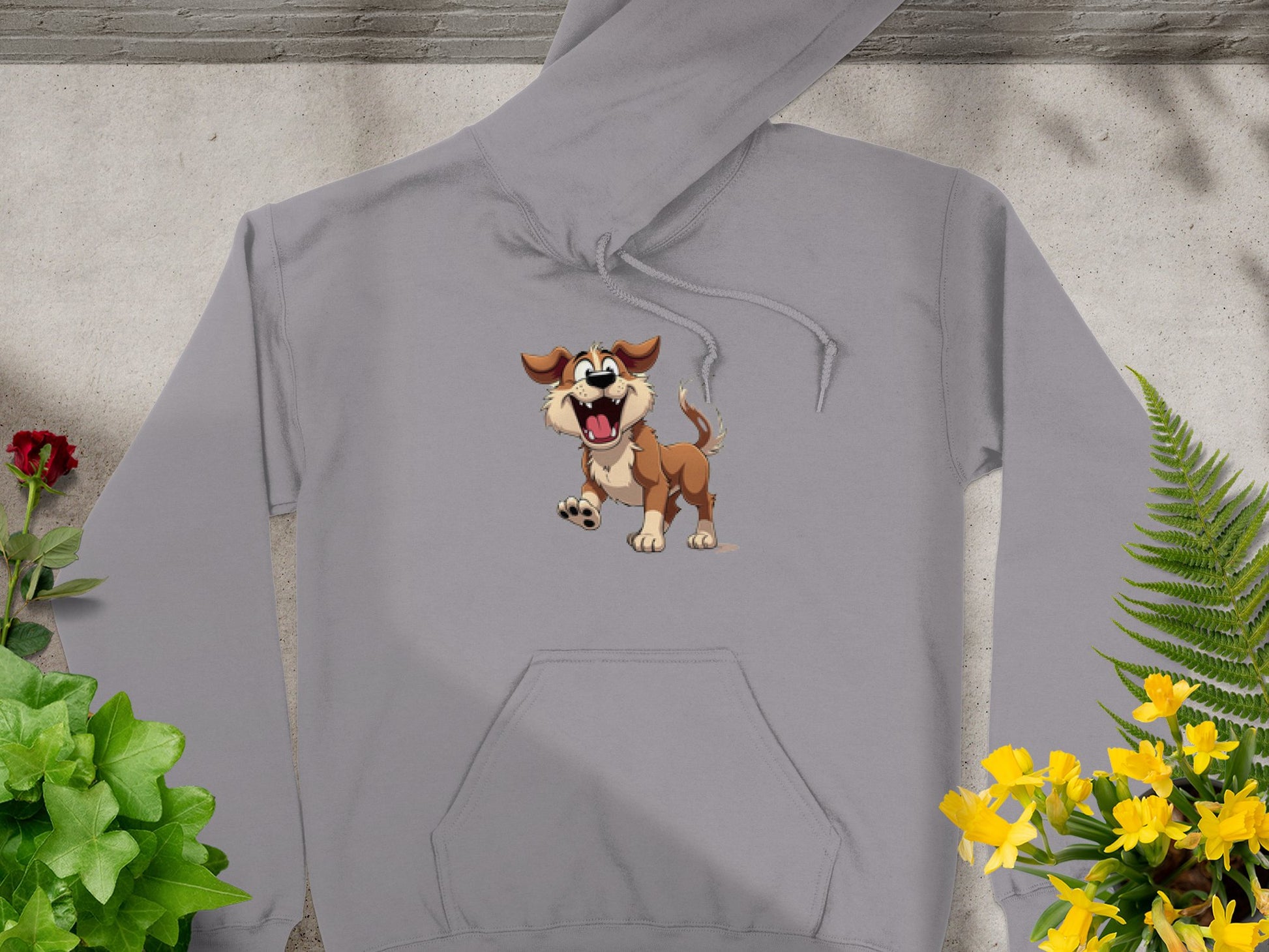 The gray hoodie from Garment Graphics, perfect for dog enthusiasts, features a cartoon of a joyful brown and white dog with an open mouth. Displayed on concrete amidst vibrant greenery and colorful flowers, its a fun design sure to catch the eye!.