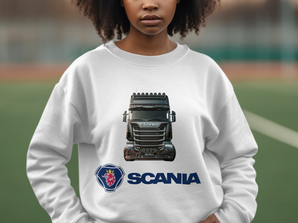 A person wears a Garment Graphics white crewneck sweatshirt featuring a striking image of a black Scania truck with the logo and Scania name below. The background is blurred, outdoors, appealing to truck enthusiasts.