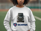 A person wears a Garment Graphics white crewneck sweatshirt featuring a striking image of a black Scania truck with the logo and Scania name below. The background is blurred, outdoors, appealing to truck enthusiasts.