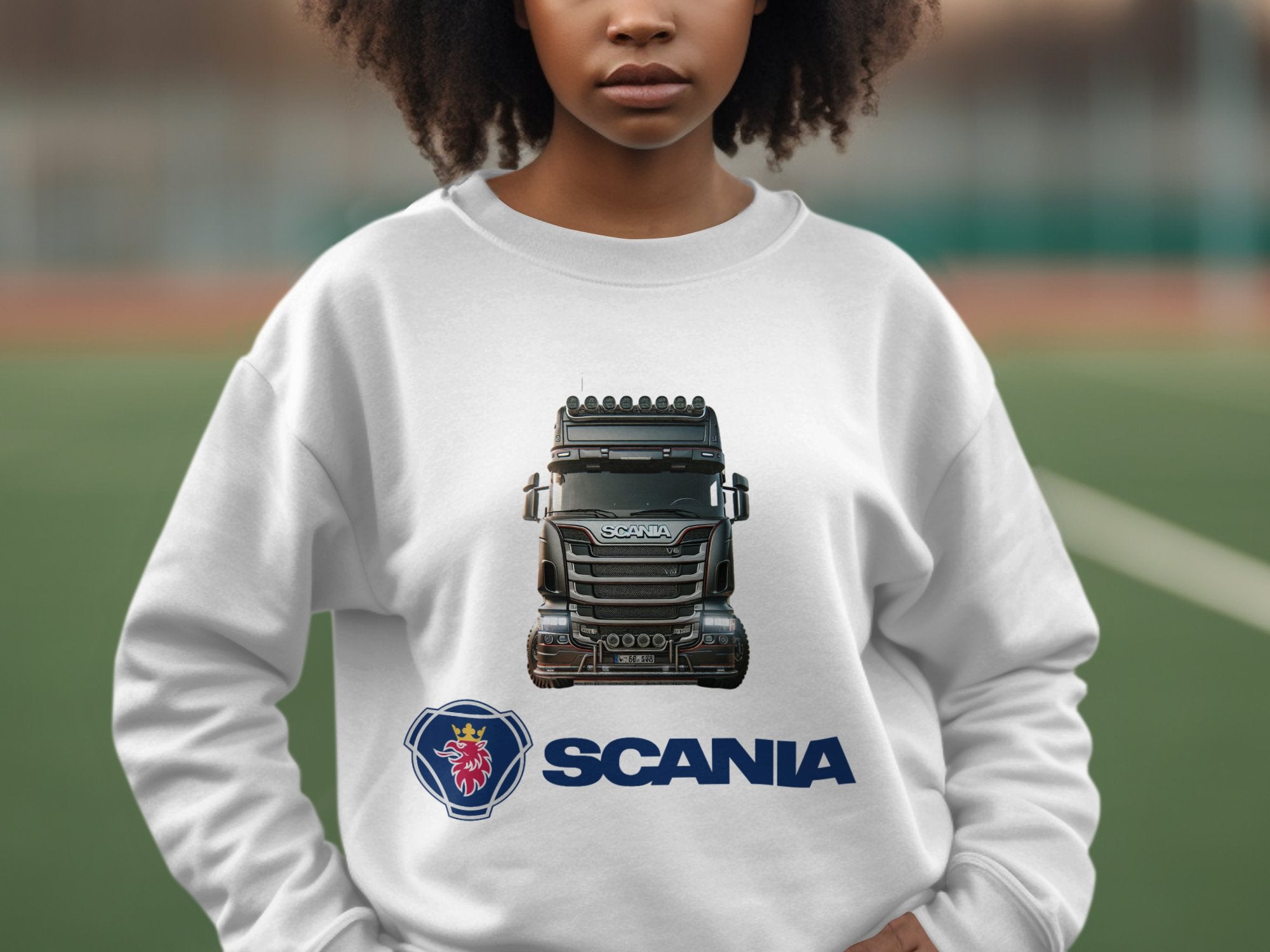 A person wears a Garment Graphics white crewneck sweatshirt featuring a striking image of a black Scania truck with the logo and Scania name below. The background is blurred, outdoors, appealing to truck enthusiasts.