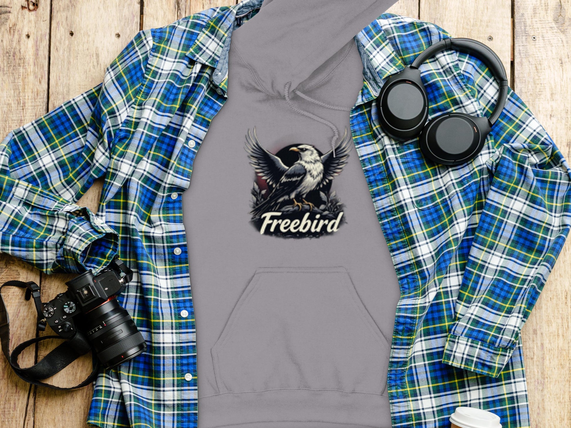 A Garment Graphics gray Freebird Graphic Pullover Hoodie featuring a bird design is displayed on a blue and green flannel shirt atop a wooden surface. Nearby, youll find a camera, headphones, and a white travel mug, highlighting your distinctive style.