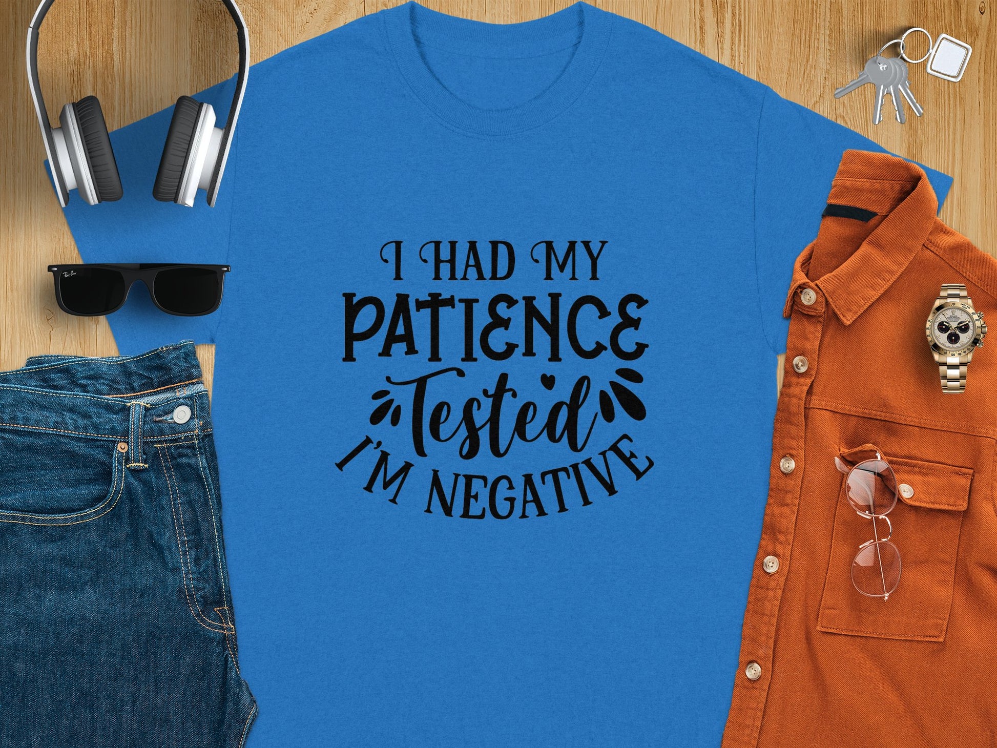 The Garment Graphics blue t-shirt, featuring the humorous phrase I Had My Patience Tested, Im Negative in black text, is styled with accessories like headphones, sunglasses, blue jeans, a burnt orange shirt, a watch, and keys on a wooden surface—ideal for expressing your sarcastic attitude.