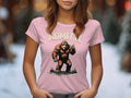 A woman wears a pink Garment Graphics T-shirt featuring a cartoon Bigfoot flashing a peace sign with the words ADMIT IT BIGFOOT. LIFE WOULD BE BORING WITHOUT ME, standing out vividly against a blurred outdoor background.