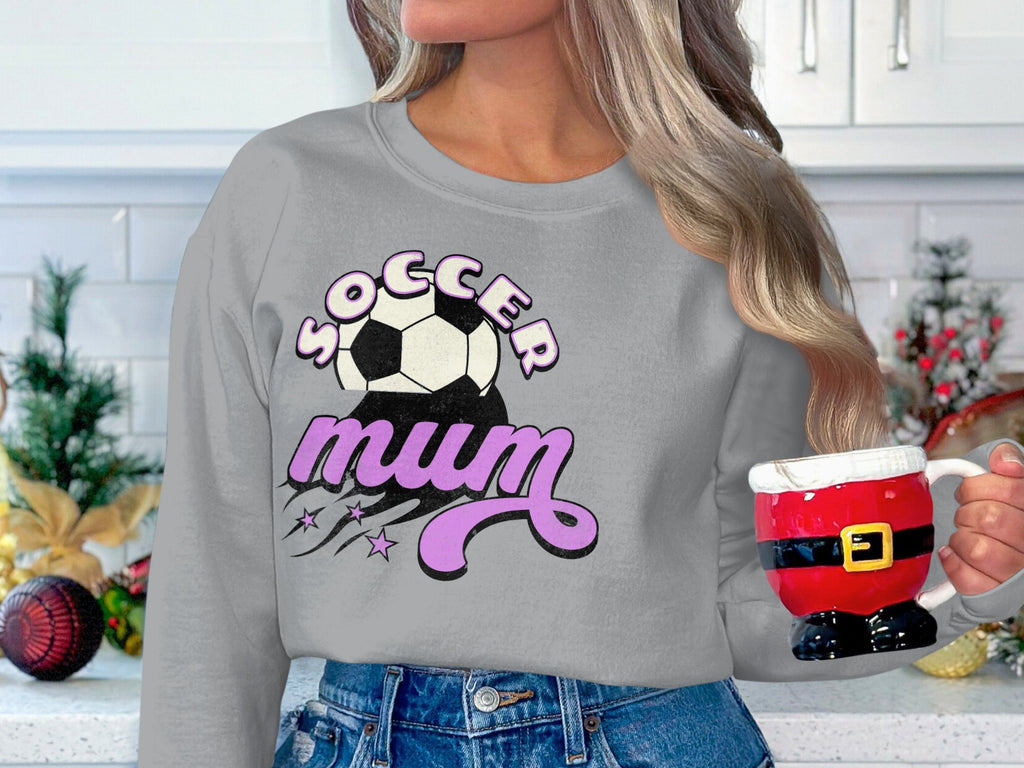 A person wearing a vintage-style Garment Graphics gray sweatshirt with the Soccer Mum graphic and soccer ball holds a Santa-themed mug shaped like Santas pants, set against a festively decorated kitchen backdrop.