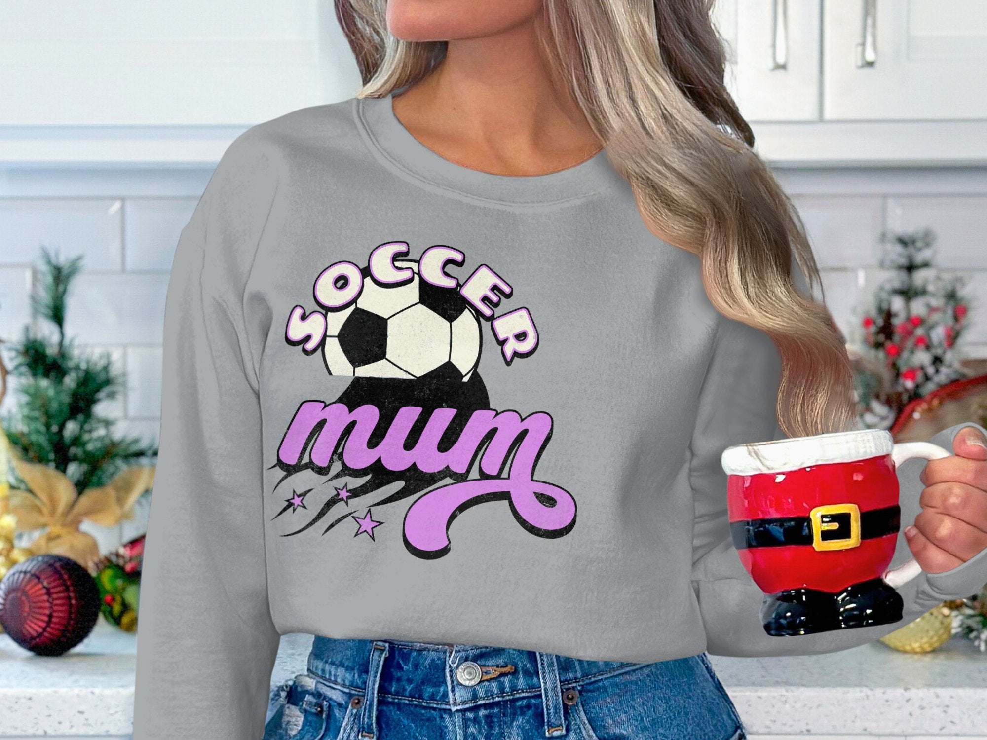 A person wearing a vintage-style Garment Graphics gray sweatshirt with the Soccer Mum graphic and soccer ball holds a Santa-themed mug shaped like Santas pants, set against a festively decorated kitchen backdrop.