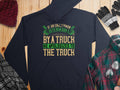 A navy hoodie from Garment Graphics sits on a wooden surface with a maroon cap, gloves, a plaid shirt, and a checked scarf. It features the humorous quote: If an Englishman gets run over by a truck, he apologizes to the truck, showcasing classic English etiquette.