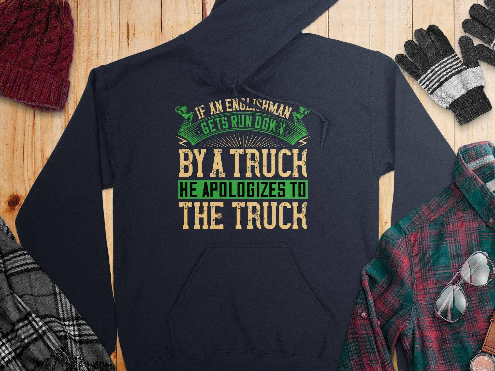 A navy hoodie from Garment Graphics sits on a wooden surface with a maroon cap, gloves, a plaid shirt, and a checked scarf. It features the humorous quote: If an Englishman gets run over by a truck, he apologizes to the truck, showcasing classic English etiquette.